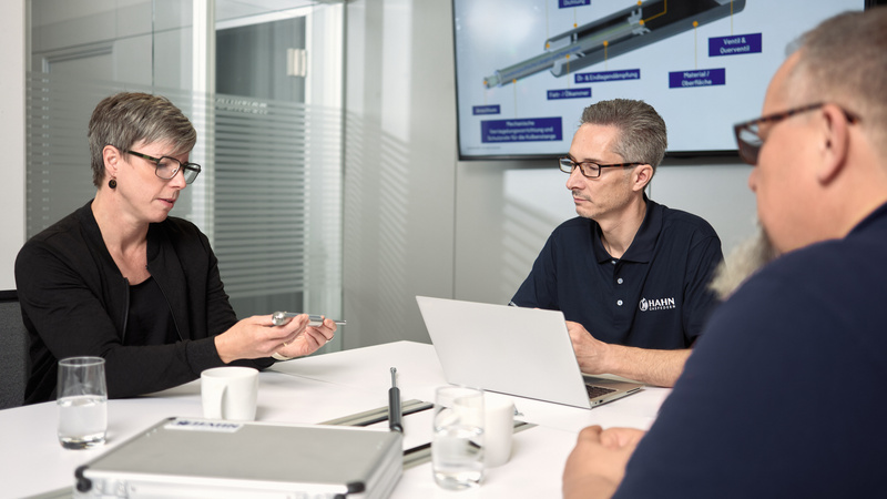 Technisches Meeting bei Hahn Gasfedern: Eine Mitarbeiterin zeigt eine Gasdruckfeder, während zwei Mitarbeiter aufmerksam zuhören und diskutieren.
