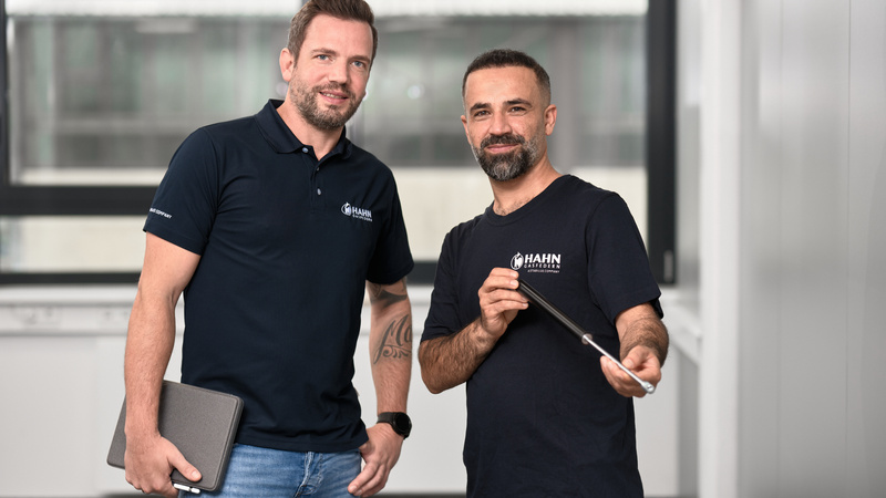 Zwei Mitarbeitende von Hahn Gasfedern stehen lächelnd zusammen, einer hält eine Gasdruckfeder, der andere ein Laptop – Teamarbeit im Fokus.