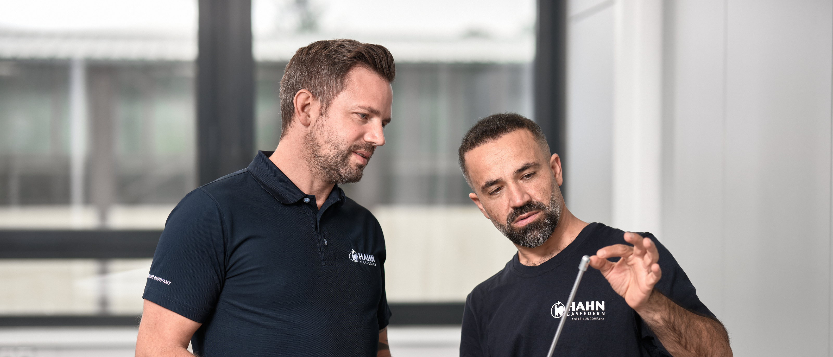 Zwei Mitarbeiter von Hahn Gasfedern begutachten eine hochwertige Gasdruckfeder und prüfen dessen Präzision und Qualität in einem modernen Arbeitsumfeld.