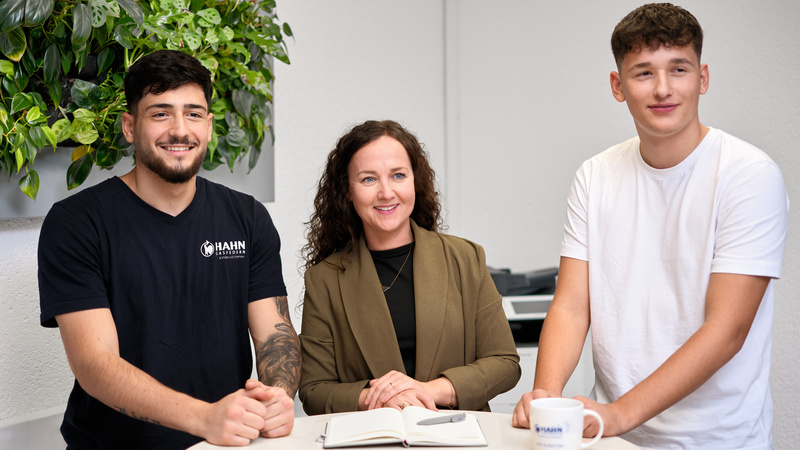 Zwei Auszubildende und Ihre Ausbilderin von Hahn Gasfedern lächeln in die Kamera, stehen an einem Tisch mit Notizbuch und Kaffeetasse – Teamwork und Austausch.