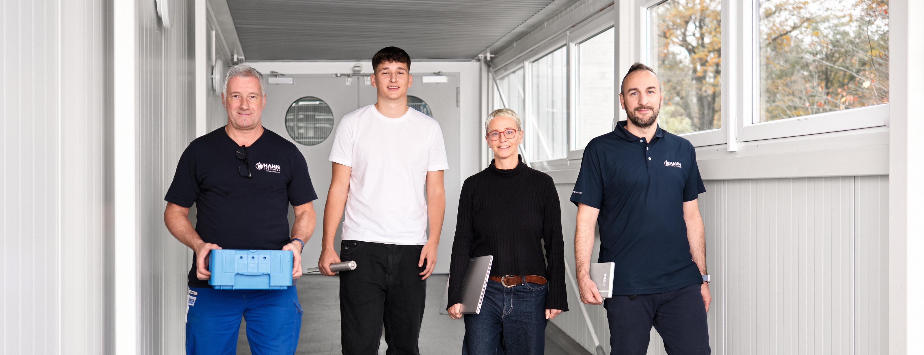 Vier Mitarbeitende von Hahn Gasfedern aus verschiedenen Unternehmensbereichen gehen gemeinsam einen lichtdurchfluteten Gang entlang, symbolisch für Teamarbeit und Innovation.