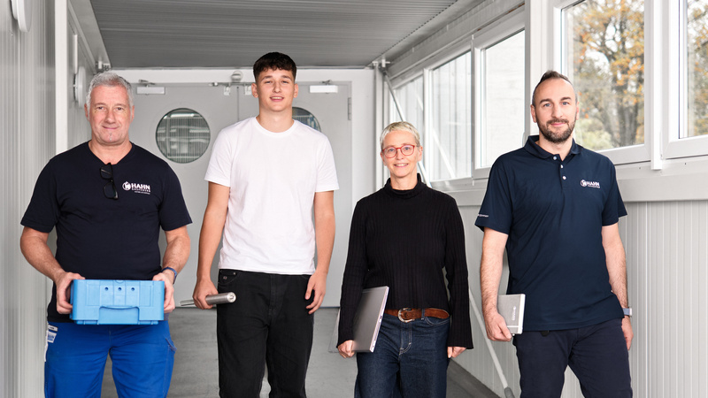 Vier Mitarbeitende von Hahn Gasfedern aus verschiedenen Unternehmensbereichen gehen gemeinsam einen lichtdurchfluteten Gang entlang, symbolisch für Teamarbeit und Innovation.