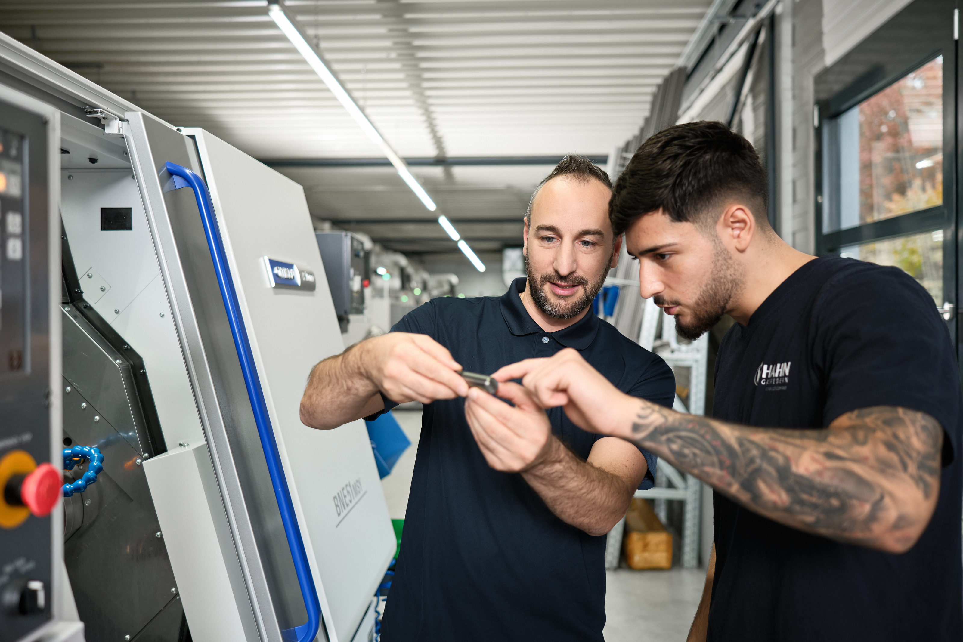 Mitarbeiter bei Hahn Gasfedern erklärt einem Auszubildenden ein Bauteil an einer modernen CNC-Maschine, praxisnahe Ausbildung in der Fertigungstechnik.