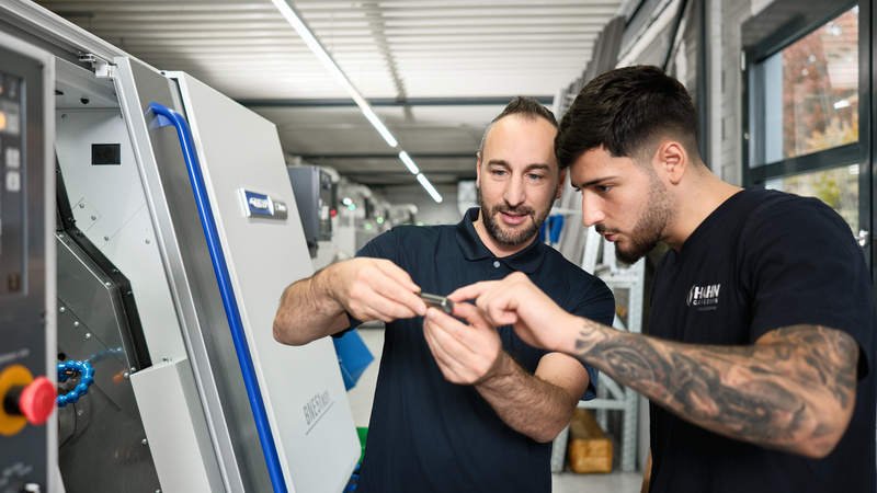 Mitarbeiter bei Hahn Gasfedern erklärt einem Auszubildenden ein Bauteil an einer modernen CNC-Maschine, praxisnahe Ausbildung in der Fertigungstechnik.