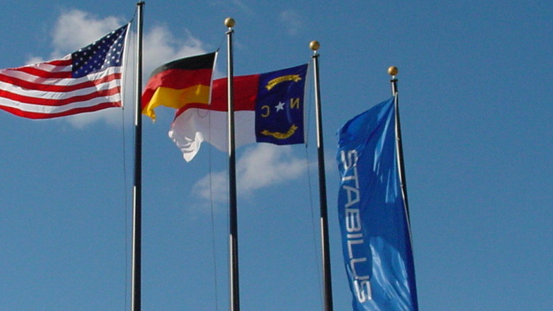 Vier Fahnenmasten vor einem Gebäude mit Stabilus-Logo: die Flaggen der USA, Deutschland, North Carolina und Stabilus – Symbol für die internationale Präsenz von HAHN-Gasfedern und die Verbindung zwischen Deutschland und den USA.
