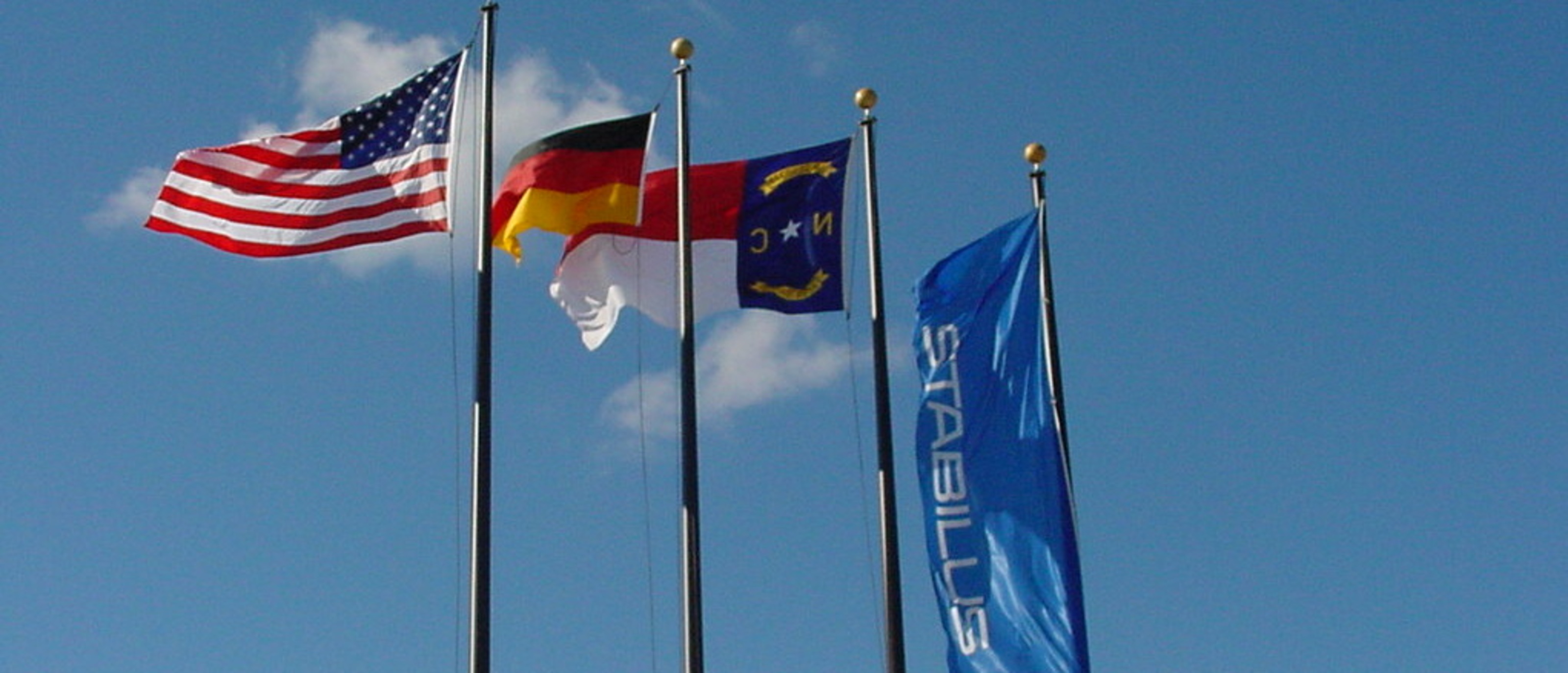 Vier Fahnenmasten vor einem Gebäude mit Stabilus-Logo: die Flaggen der USA, Deutschland, North Carolina und Stabilus – Symbol für die internationale Präsenz von HAHN-Gasfedern und die Verbindung zwischen Deutschland und den USA.