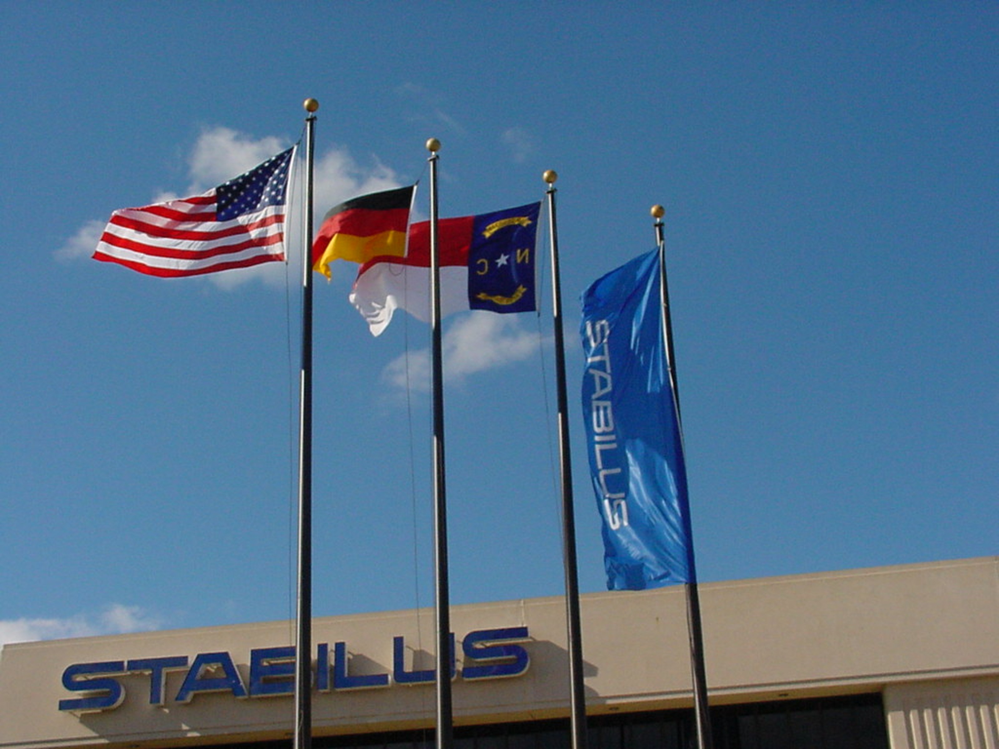 Vier Fahnenmasten vor einem Gebäude mit Stabilus-Logo: die Flaggen der USA, Deutschland, North Carolina und Stabilus – Symbol für die internationale Präsenz von HAHN-Gasfedern und die Verbindung zwischen Deutschland und den USA.